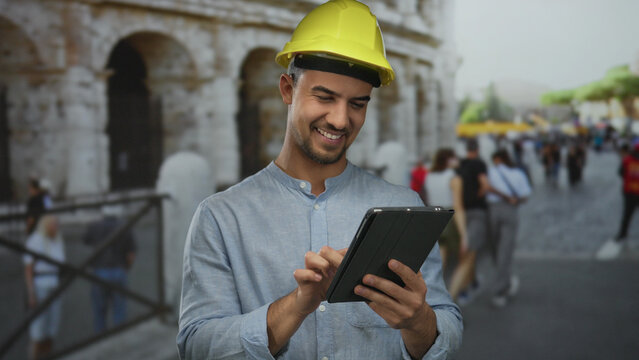 Fototapeta Young hispanic man in a hardhat using a tablet outside the roman coliseum, surrounded by city street activity, conveying an urban work environment in rome, italy.