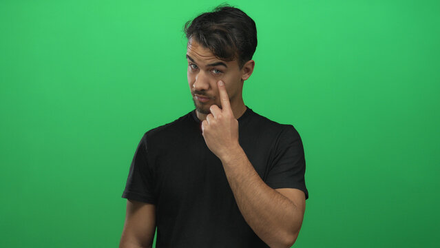 Young hispanic man in black shirt gesturing vigilance over green background displaying serious facial expression with isolated focus - Powered by Adobe