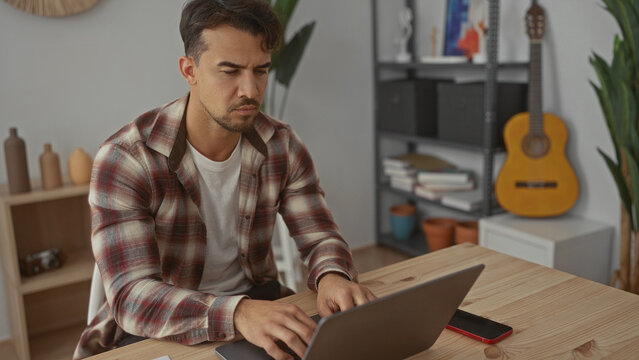 Young hispanic man working on a laptop in a modern office setting with a guitar and shelves in the background, conveying a focused and casual work atmosphere.