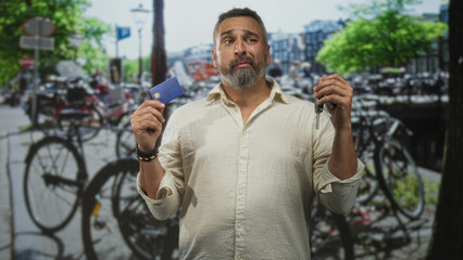 Middleaged man holding creditcard and keys with both hands on street in amsterdam; indecision travel planning.