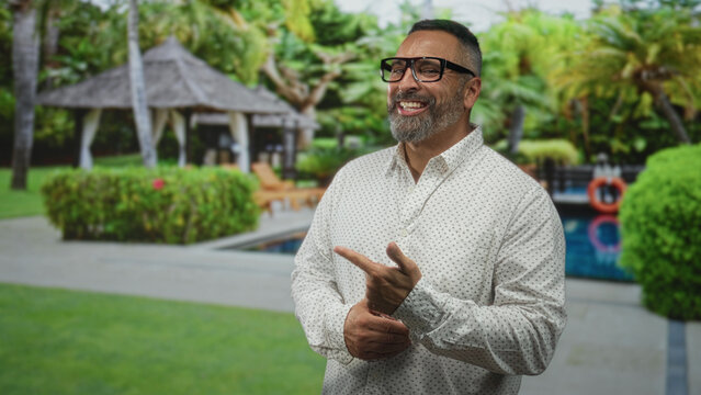 Man with beard and grey hair pointing with hand near poolside cabana by outdoor pool, wearing glasses and patterned shirt; cheerful.
