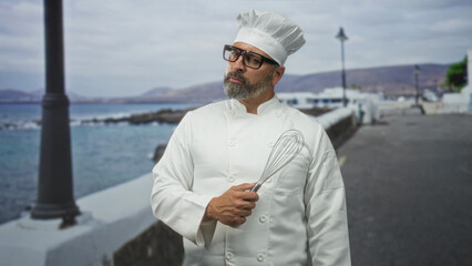 Man chef wearing white uniform and toque holds metal whisk in right hand on seaside promenade by ocean wall; pride tradition craft.