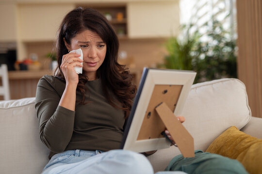Emotional young woman wiping tears while looking at a framed photo, expressing sadness, longing and emotional pain.
