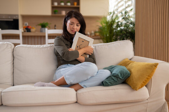 Young woman sitting on a sofa and hugging a framed photo with a sad, emotional expression at home.
