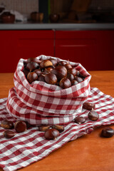 Fresh chestnuts displayed in a red checkered cloth bag on a wooden kitchen table, with scattered chestnuts creating a warm and rustic seasonal atmosphere.