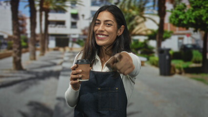 Woman points finger at viewer while holding takeaway coffee cup wearing denim apron on street; welcoming local cafe.