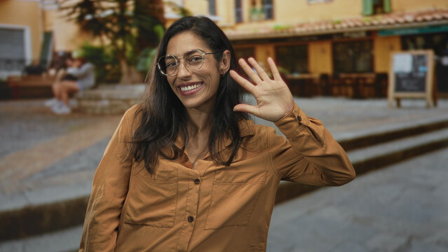 Woman in brown shirt smiling and waving her right hand on a stone street with cafe steps and outdoor terrace; friendly greeting.
