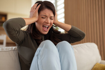 A woman sitting on a sofa, holding her head and shouting in frustration, expressing strong...