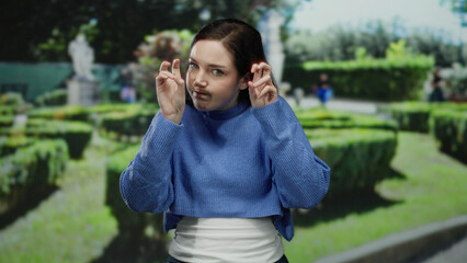 Brunette woman in blue sweater gestures humorously in sunny park with lush greenery and blurred hedge maze, suggesting playful interaction and a lighthearted outdoor atmosphere.
