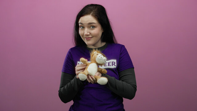 Woman embracing stuffed animal against vivid pink background wearing volunteer shirt highlighting tenderness and compassion in humanitarian context - Powered by Adobe