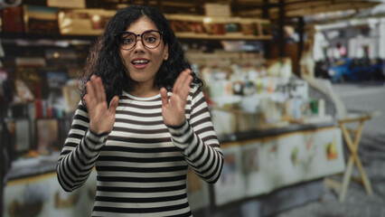 Woman with hands pressed together at street market stall, wearing glasses and black and white striped top, slight smile at outdoor souvenir stand; gratitude hope.