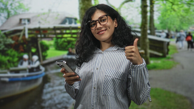Young arab woman holding smartphone and giving thumbs up on a street by a canal and wooden bridge; confidence connection. - Powered by Adobe