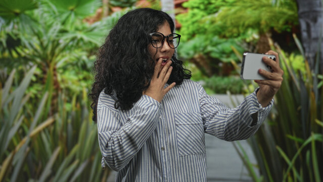 Young arab woman holding smartphone and taking a selfie in a park pathway wearing striped shirt and glasses; casual surprise.