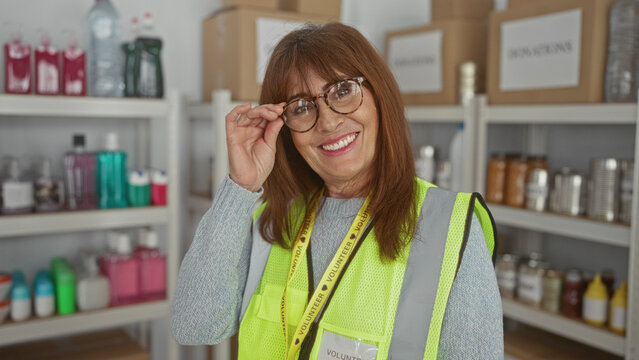 Senior hispanic woman volunteering at an indoor donation center wearing a reflective vest and smiling among shelves filled with various charity supplies.