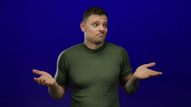 Young man with tattooed arm in green shirt shrugging and expressing confusion against blue background