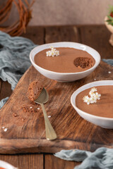 Individual servings of smooth chocolate mousse in white bowls, one with a spoonful lifted, garnished with tiny white flowers, arranged on a rustic wooden board