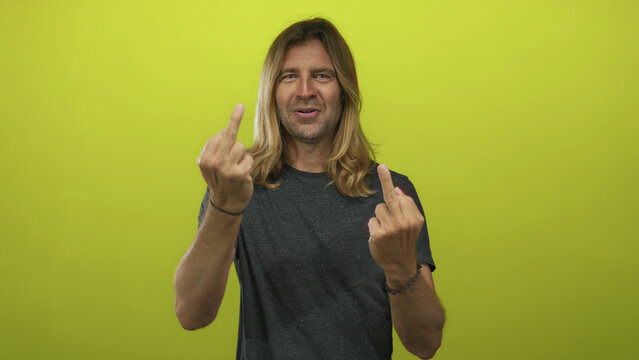 Blond man with long hair sticks tongue out and flips both middle fingers in a studio setting; defiance.