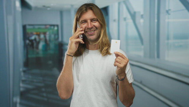 Man smiling broadly while speaking on smartphone and holding creditcard at an airport terminal; happiness.