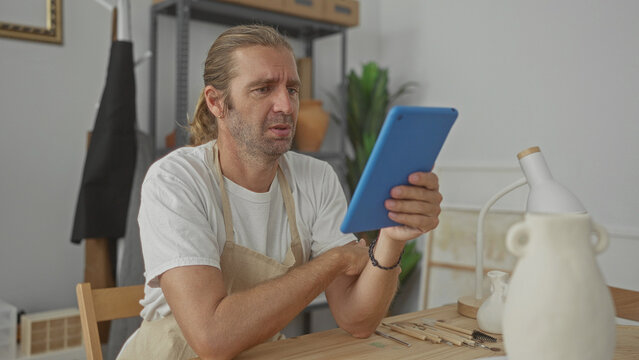 Man holds blue tablet in pottery studio while wearing apron and examining ceramic sketches; creative focus.