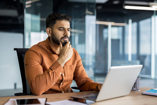 Thoughtful indian businessman focusing intently, organizing new ideas and contemplating solutions while working on his laptop at a wooden desk in a contemporary corporate office environment