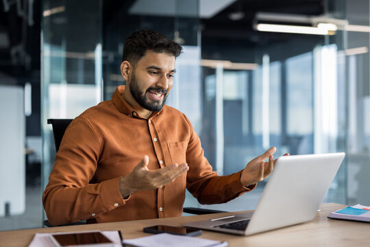 Indian businessman smiling and talking during a video conference on his laptop, actively engaging in a virtual business meeting inside a modern office workspace