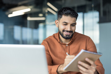 Fototapeta premium Young businessman smiling, making notes in a notebook while working on a laptop in a modern office, focusing on his tasks and professional development