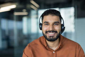 Young indian customer service agent with headset smiling confidently in modern office, providing...