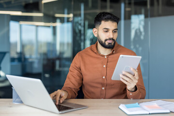 Young businessman working remotely on a laptop and holding a digital tablet, demonstrating productivity and efficient use of technology in a modern office environment