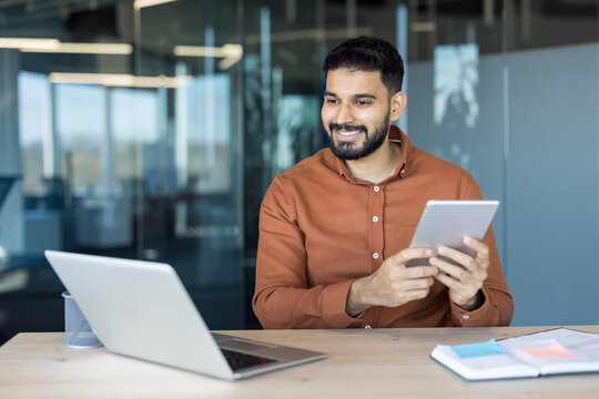 Young businessman participating in a virtual meeting or online presentation, holding a digital tablet and smiling while sitting at his desk with a laptop in a modern office environment