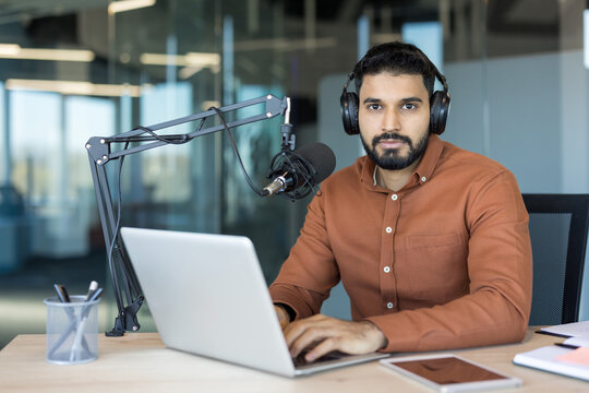 Young man with headphones and microphone typing on laptop in a modern office, recording and live-streaming podcast audio content from a professional home studio workspace