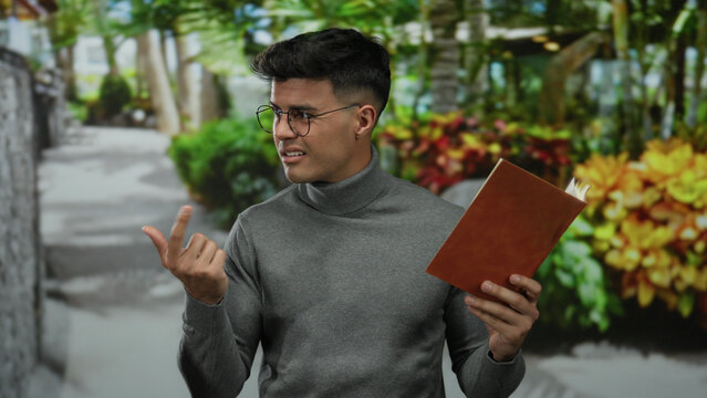 Young man reading book outdoors in park surrounded by lush greenery while wearing glasses and sweater in thoughtful expression.