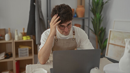 Man with hand to head over laptop at studio table, wearing apron and glasses, leaning forward among pottery and shelves; frustration creative block.