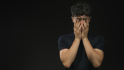 Young man with hands covering face in black studio wearing glasses with closed eyes and tense posture; anxiety isolation introspection.