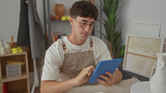 Man tapping tablet with fingers in studio while wearing apron and pottery nearby; concentration craftsmanship.