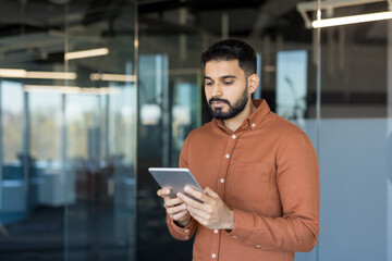 Indian businessman in a brown shirt standing in a modern office, focused on a tablet while analyzing data and using apps for communication and productivity