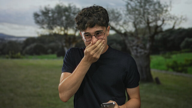 Man with hand covering mouth and holding smartphone in forest field near trees while looking at screen; shock privacy concern.