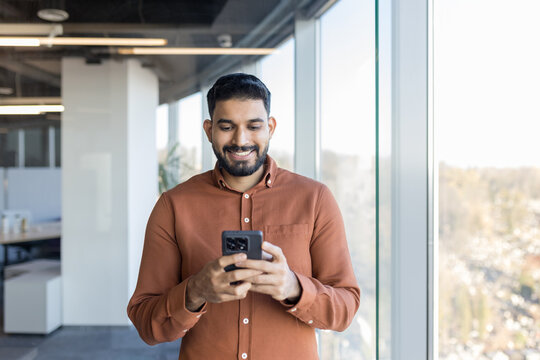 Indian businessman smiling while checking messages on his smartphone, connecting with people, and managing business in a contemporary office building with large windows