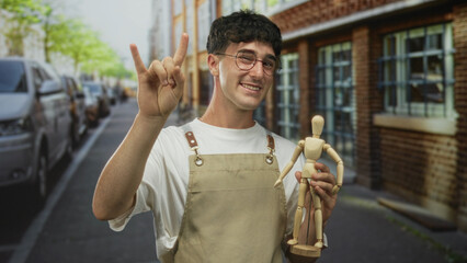 Young man wearing apron and tshirt holding a wooden mannequin and making rock horns with his hand on a city street; creative craft playful.