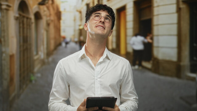 Man in white shirt holding tablet looks up on a cobbled city street between old buildings, wearing glasses and intent expression; curiosity. - Powered by Adobe