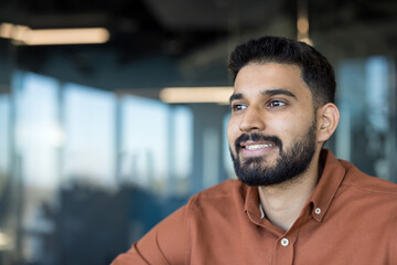 Young adult indian man posing for a half body portrait, confidently smiling and looking away, showcasing professionalism and a positive outlook in a modern office environment
