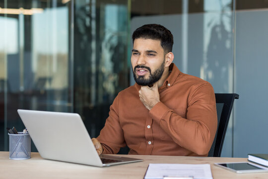 Young man rubbing his sore throat during a virtual meeting, looking unwell and uncomfortable while working remotely from a modern office desk with laptop and headset