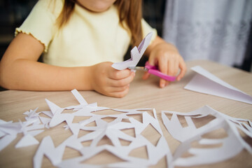 A child creating paper white snowflakes at a table at home. A child cuts patterns on paper with scissors. Decoration for the winter holiday.