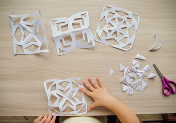 A child creating paper white snowflakes at a table at home. A child cuts patterns on paper with scissors. Decoration for the winter holiday.