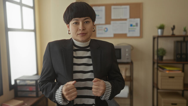 Woman standing confidently in an office with arms folded, wearing a striped sweater and black blazer, surrounded by office decor and charts on the wall, light streaming in from window.
