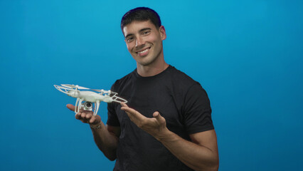 Young hispanic man holds a white drone in one hand and points finger to object in studio; curiosity.