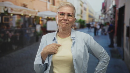Woman finger pointing to chest on busy street terrace, short cropped haircut, eyeglasses, light shirt, hand on hip, direct gaze; confidence.