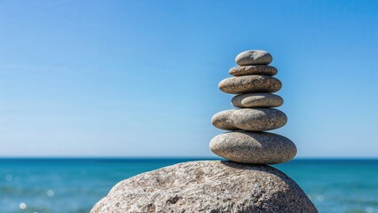 Fototapeta premium Stacked stones on a large rock by the sea with a clear blue sky. Balance and serenity, nature, and meditation.