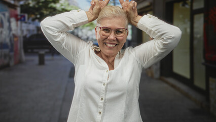 Woman middle aged smiling with hands making peace signs above head on city street wearing glasses and white button shirt short hair visible; playful joy.