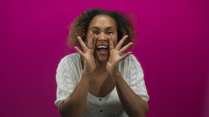 Woman with visible fingers and open mouth cupping hands to shout wearing a white knit top in a bright pink studio; joy.
