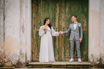 Bride in elegant white dress holding bouquet stands beside groom in gray suit, both smiling in front of weathered green doors, capturing a joyful wedding moment
